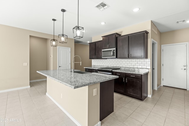 kitchen featuring light stone counters, decorative backsplash, a center island with sink, dark brown cabinets, and appliances with stainless steel finishes