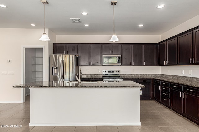 kitchen featuring appliances with stainless steel finishes, dark brown cabinets, hanging light fixtures, dark stone countertops, and a kitchen island with sink