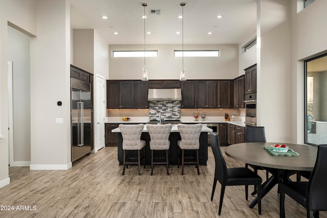 kitchen with light wood-type flooring, hanging light fixtures, appliances with stainless steel finishes, exhaust hood, and a high ceiling