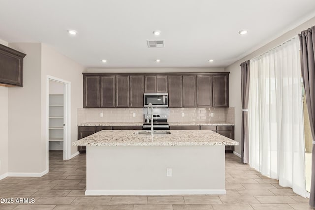 kitchen with dark brown cabinetry, sink, light stone counters, a center island with sink, and appliances with stainless steel finishes