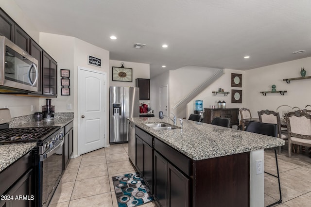 kitchen featuring appliances with stainless steel finishes, light tile patterned floors, dark brown cabinets, a kitchen island with sink, and sink