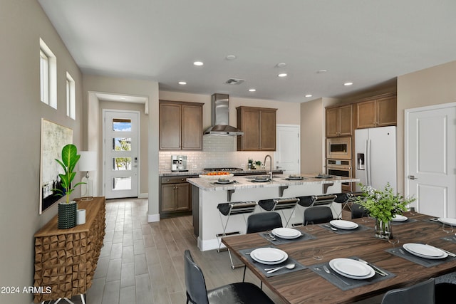 kitchen featuring light stone countertops, a kitchen island with sink, wall chimney range hood, a breakfast bar area, and appliances with stainless steel finishes