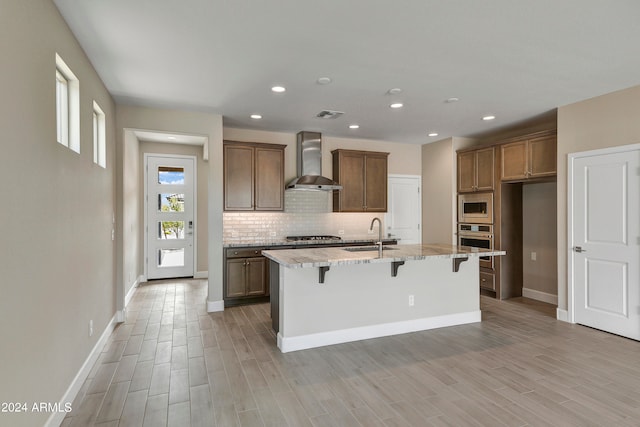 kitchen with wall chimney range hood, light wood-type flooring, a kitchen breakfast bar, stainless steel appliances, and an island with sink