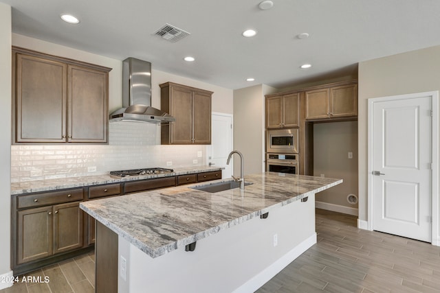 kitchen featuring sink, a center island with sink, wall chimney exhaust hood, stainless steel appliances, and light stone countertops