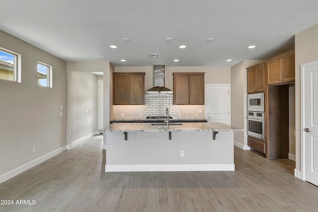 kitchen featuring a center island with sink, wall chimney exhaust hood, appliances with stainless steel finishes, a breakfast bar, and light stone countertops