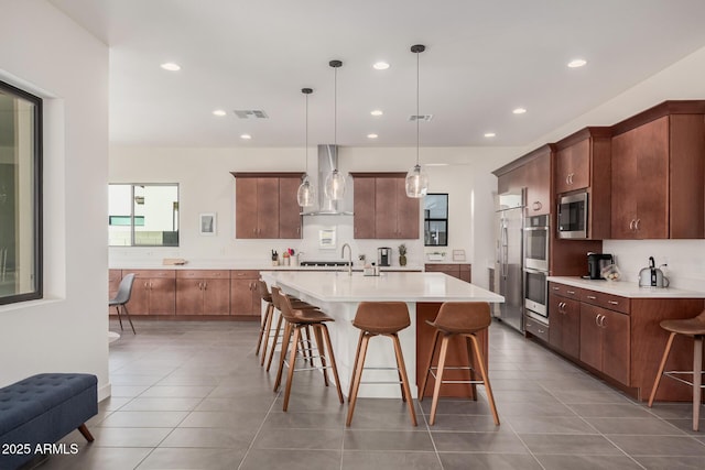 kitchen featuring appliances with stainless steel finishes, an island with sink, a kitchen breakfast bar, hanging light fixtures, and wall chimney exhaust hood