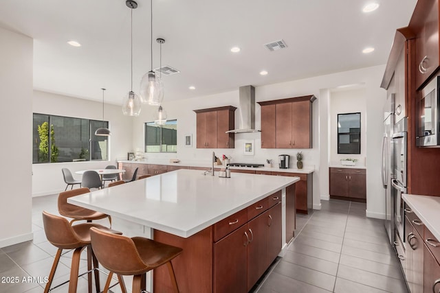 kitchen with pendant lighting, wall chimney range hood, appliances with stainless steel finishes, a kitchen breakfast bar, and an island with sink