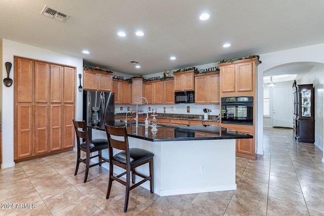 kitchen with a kitchen island with sink, dark stone counters, a kitchen breakfast bar, black appliances, and sink