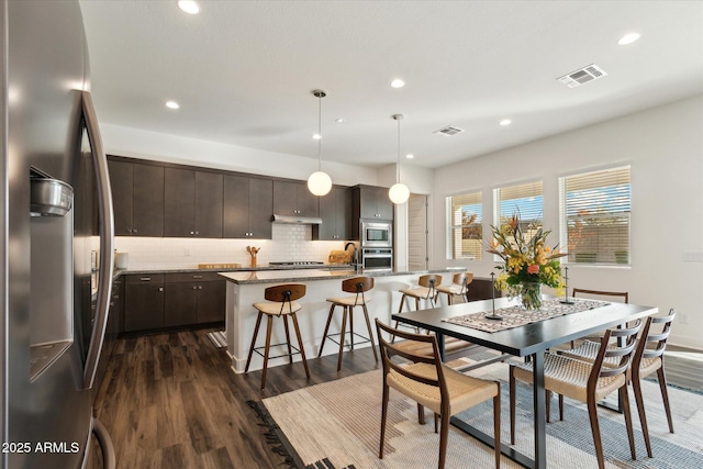 kitchen featuring pendant lighting, dark brown cabinets, stainless steel appliances, a center island with sink, and dark hardwood / wood-style flooring