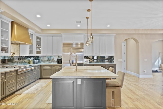 kitchen with wall chimney exhaust hood, an island with sink, decorative light fixtures, gray cabinets, and appliances with stainless steel finishes