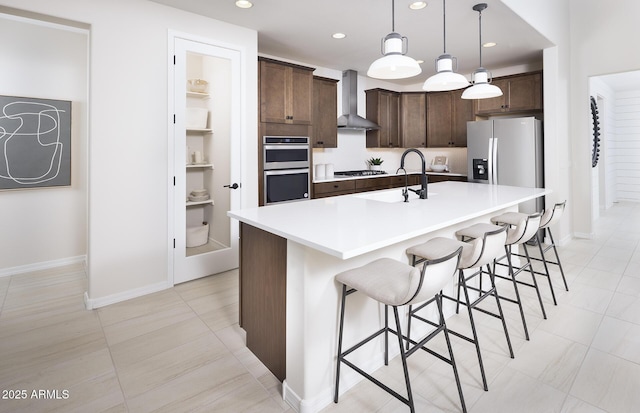 kitchen featuring pendant lighting, appliances with stainless steel finishes, dark brown cabinets, a center island with sink, and wall chimney exhaust hood