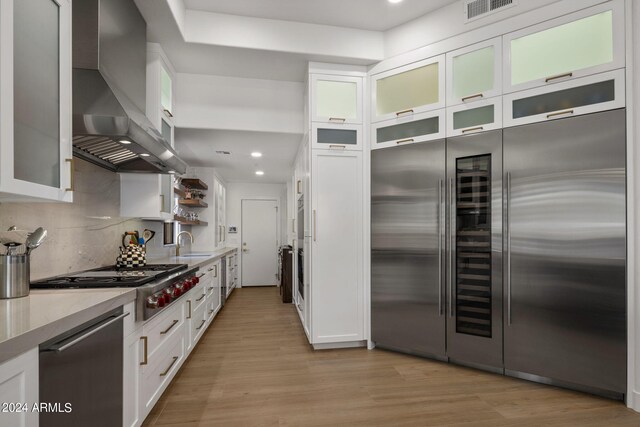 kitchen with stainless steel appliances, sink, wall chimney range hood, white cabinetry, and light hardwood / wood-style flooring