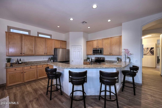 kitchen with a kitchen bar, dark wood-type flooring, appliances with stainless steel finishes, light stone counters, and kitchen peninsula