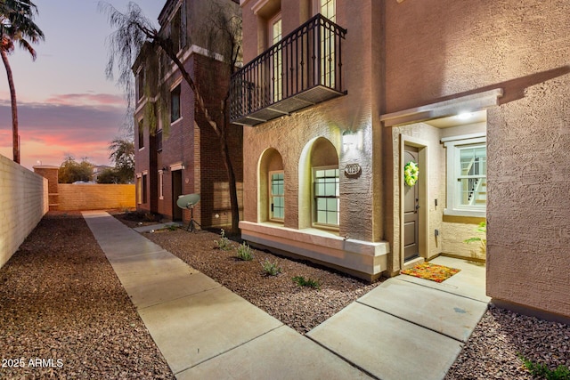 property exterior at dusk featuring fence, a balcony, and stucco siding