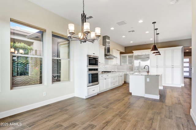 kitchen with wall chimney exhaust hood, white cabinetry, built in appliances, decorative light fixtures, and a center island with sink