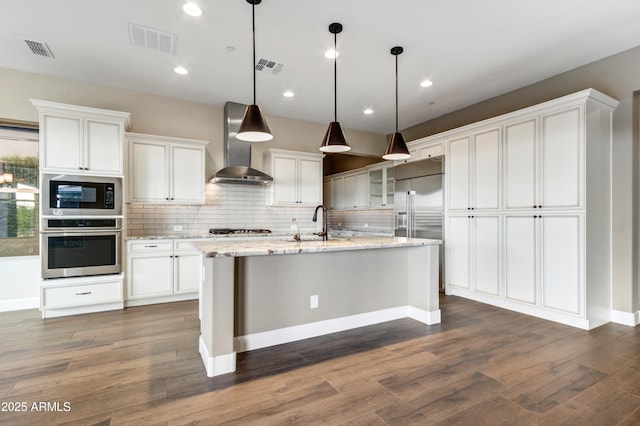 kitchen featuring wall chimney range hood, white cabinetry, hanging light fixtures, built in appliances, and light stone counters