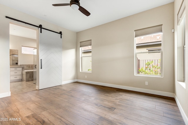 unfurnished bedroom featuring ceiling fan, ensuite bath, a barn door, and light hardwood / wood-style flooring