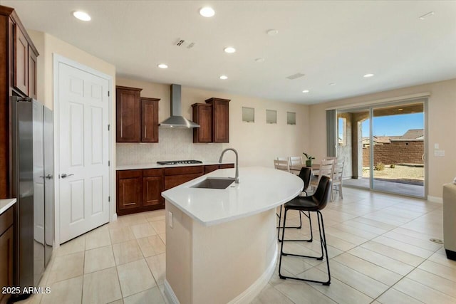 kitchen featuring sink, wall chimney range hood, stainless steel fridge, a breakfast bar, and a center island with sink