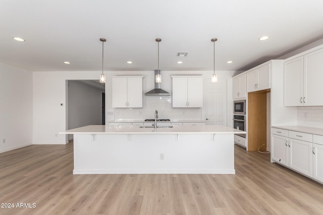 kitchen with a kitchen island with sink, stainless steel appliances, wall chimney range hood, decorative backsplash, and light wood-type flooring