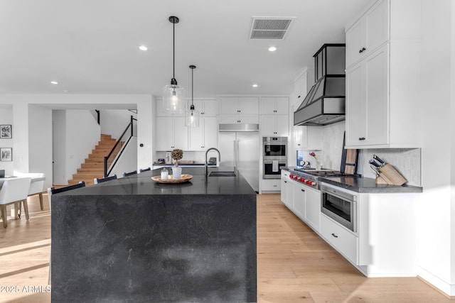 kitchen with tasteful backsplash, a kitchen island with sink, built in appliances, white cabinets, and hanging light fixtures