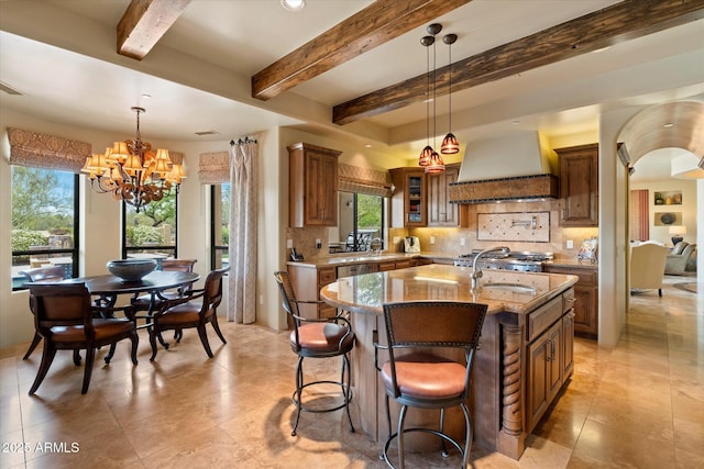 kitchen featuring tasteful backsplash, light stone countertops, a center island with sink, premium range hood, and a chandelier