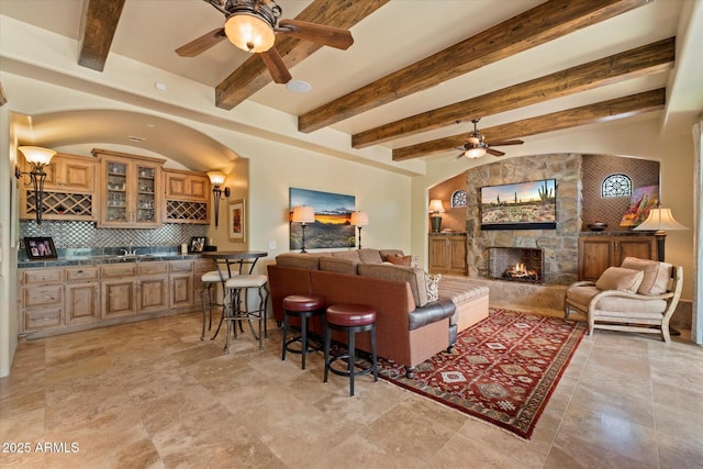 living area with indoor wet bar, a ceiling fan, a stone fireplace, and beamed ceiling