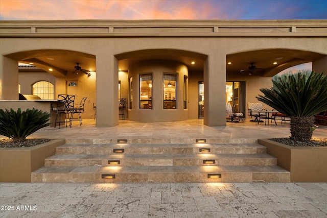 exterior entry at dusk with a ceiling fan, stucco siding, a patio area, and an outdoor bar