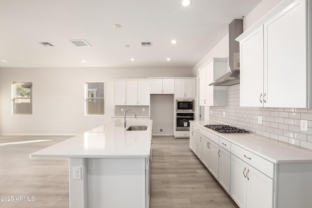 kitchen with stainless steel appliances, sink, white cabinetry, wall chimney exhaust hood, and a center island with sink