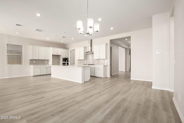 kitchen with a center island with sink, wall chimney range hood, decorative backsplash, white cabinetry, and an inviting chandelier