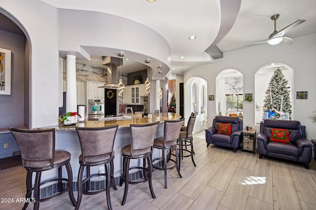 kitchen with island exhaust hood, light stone countertops, stainless steel oven, ceiling fan, and a breakfast bar area