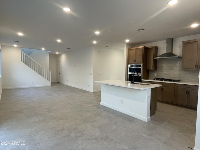 kitchen with sink, wall chimney exhaust hood, gas stovetop, decorative backsplash, and a center island with sink