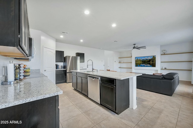 kitchen featuring sink, ceiling fan, stainless steel appliances, light stone countertops, and an island with sink