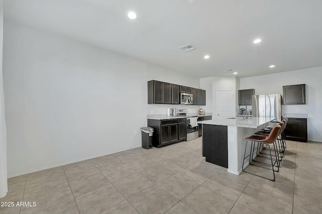 kitchen featuring sink, appliances with stainless steel finishes, a kitchen island with sink, light stone countertops, and a kitchen bar