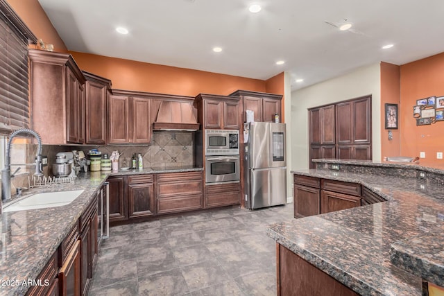 kitchen with premium range hood, a sink, backsplash, dark stone counters, and appliances with stainless steel finishes