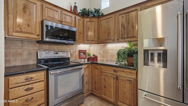 kitchen with light tile patterned flooring, stainless steel appliances, backsplash, brown cabinetry, and dark countertops