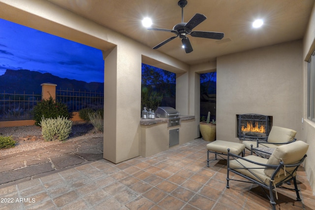 patio terrace at dusk featuring ceiling fan, area for grilling, exterior fireplace, and a grill