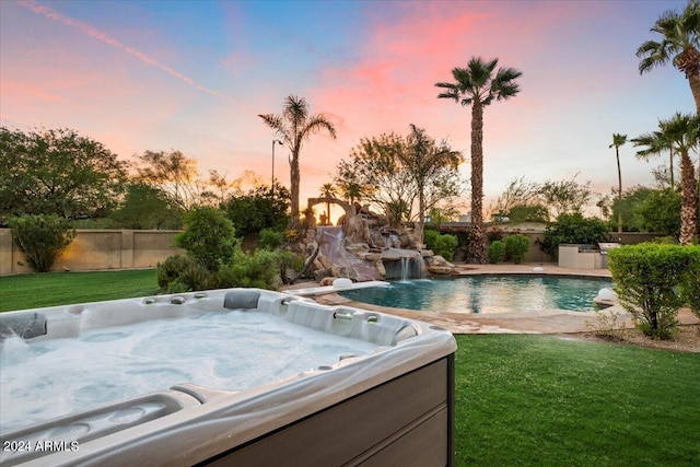 pool at dusk with a yard, pool water feature, and a hot tub