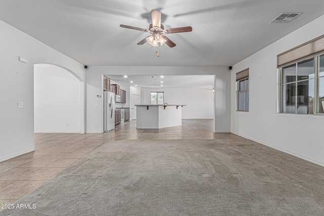 unfurnished living room with arched walkways, light tile patterned flooring, visible vents, light colored carpet, and ceiling fan