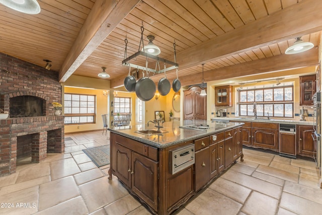 kitchen featuring a sink, a brick fireplace, beam ceiling, an island with sink, and stone tile flooring