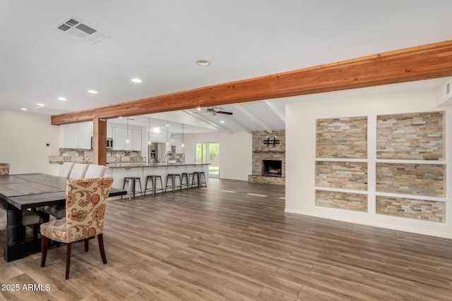 dining room with visible vents, wood finished floors, a fireplace, beamed ceiling, and a ceiling fan
