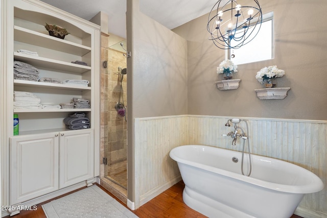 full bath featuring wood finished floors, a wainscoted wall, a freestanding tub, a stall shower, and a notable chandelier