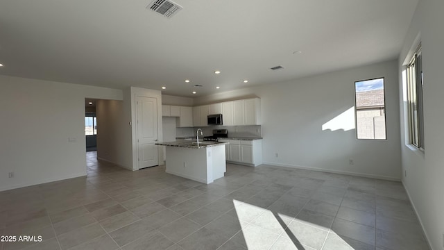 kitchen with light tile patterned floors, a kitchen island with sink, white cabinetry, stove, and dark stone counters