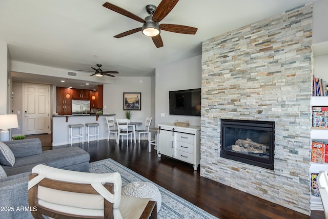 living area with dark wood-style floors, a stone fireplace, visible vents, and a ceiling fan