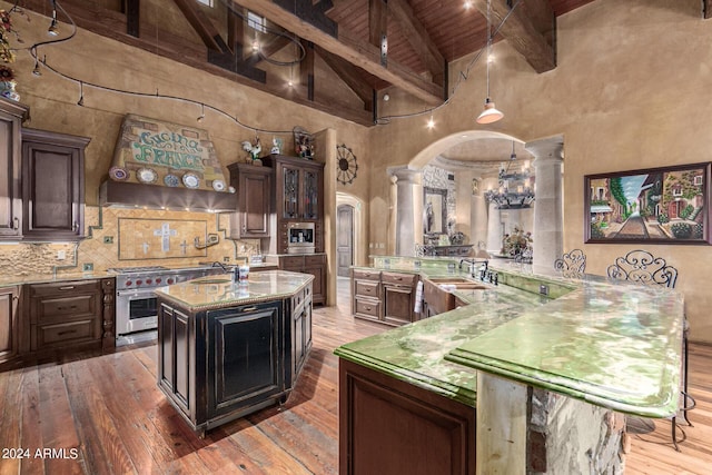 kitchen featuring high end range, ornate columns, dark brown cabinetry, a center island with sink, and beamed ceiling