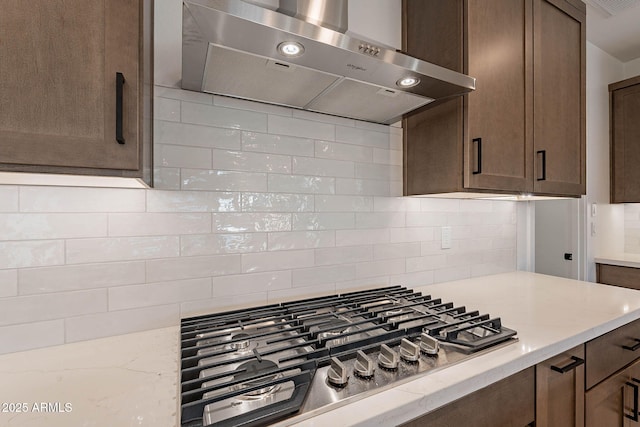 kitchen with light stone countertops, wall chimney exhaust hood, backsplash, stainless steel gas stovetop, and dark brown cabinets