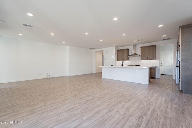 kitchen featuring light hardwood / wood-style floors, decorative backsplash, wall chimney range hood, sink, and an island with sink