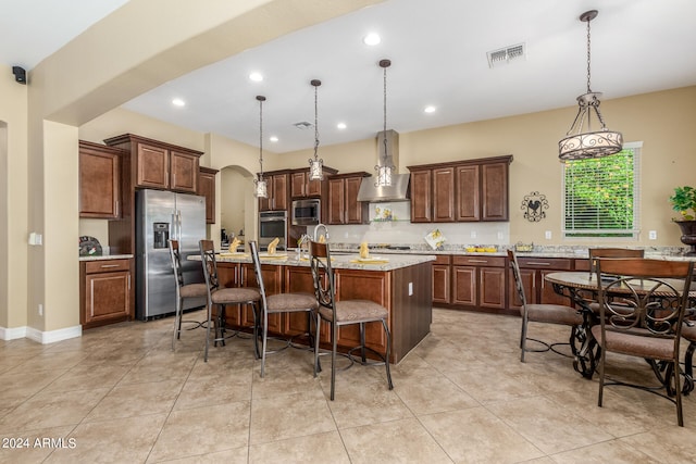 kitchen with appliances with stainless steel finishes, light stone countertops, wall chimney range hood, an island with sink, and pendant lighting