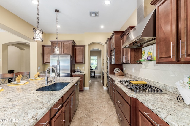 kitchen with wall chimney exhaust hood, hanging light fixtures, stainless steel appliances, sink, and light stone counters