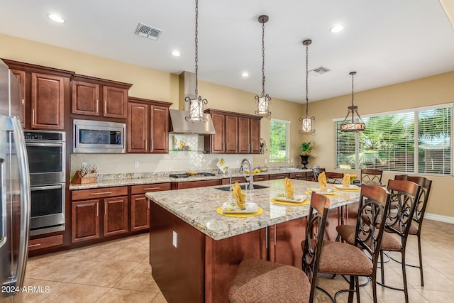 kitchen with decorative light fixtures, appliances with stainless steel finishes, light stone counters, a kitchen island with sink, and tasteful backsplash