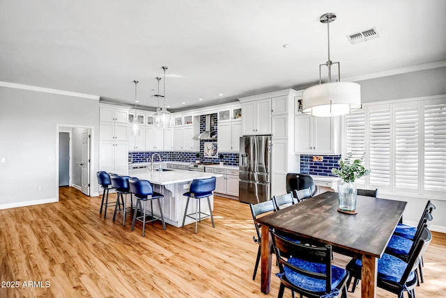 kitchen featuring wall chimney exhaust hood, stainless steel fridge, an island with sink, decorative light fixtures, and white cabinetry